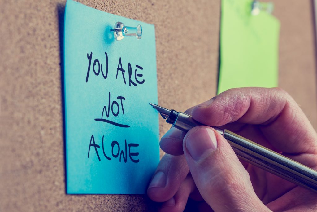 You Are Not Alone - man writing an inspirational message on a blue sticky note pinned to a cork board with a fountain pen.